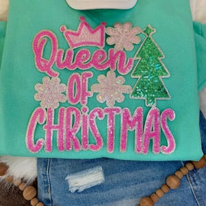 May include: A turquoise sweatshirt with the text "Queen of Christmas" in pink glitter lettering. The design includes a crown, snowflakes, and a sequined Christmas tree. A white baseball cap and blue denim shorts are also visible.