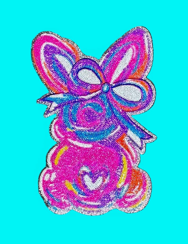 May include: A colorful, glittery bunny-shaped design with a large bow. The bunny is primarily pink with accents of blue, purple, orange, and yellow. A white heart shape is in the center of the bunny's body. The background is a solid light blue.