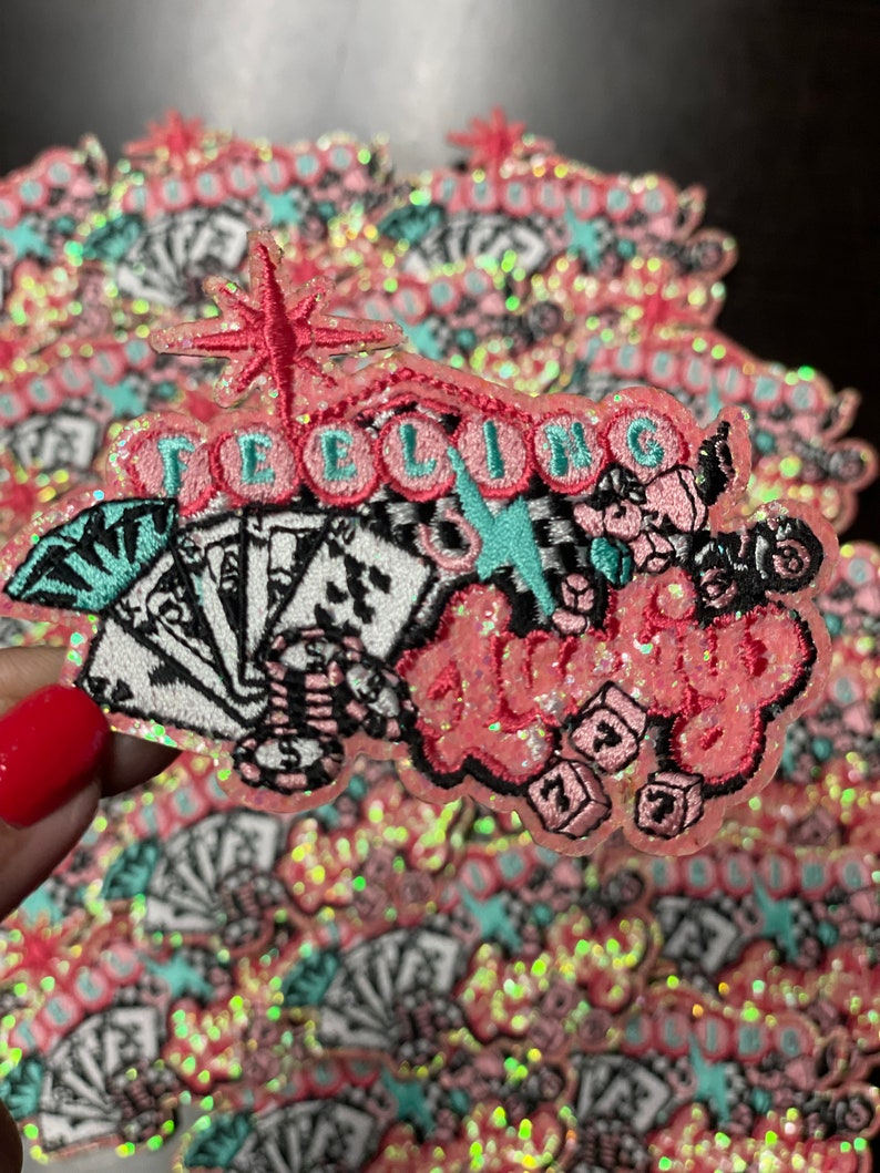 May include: Pink and glittery patch with the words "Feeling Lucky" in a cursive font. The patch features playing cards, dice, and a starburst design. The patch is surrounded by a pink border.