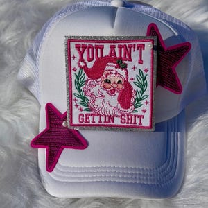 Peut inclure: Casquette de camionneur blanche avec un dos en filet et une visière incurvée. L'avant présente un écusson carré avec un Père Noël de dessin animé et le texte "YOU AIN'T GETTIN SHIT". Deux décorations en forme d'étoile rose sont attachées.