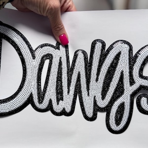 May include: A black and white sequined patch with the word "Dawgs" in a cursive font. The word is outlined in black and filled with white sequins. The patch is held against a white background.