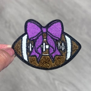 May include: Embroidered patch featuring a brown football with white stripes and a large purple bow. The patch has a black border and is likely designed for apparel or accessories, perfect for sports fans.