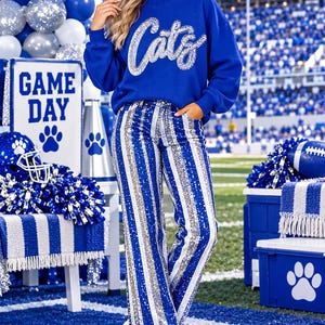 May include: A royal blue long-sleeve sweatshirt featuring the word Cats in silver sequins across the chest, paired with high-waisted pants decorated with vertical stripes of alternating royal blue and silver sequins and white platform boots.