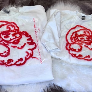 May include: Two white sweatshirts with a red Santa Claus face design. The Santa face is printed on a clear plastic sheet. The sweatshirts are on a white fur-like surface.