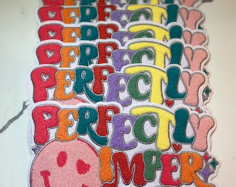 Op de afbeelding: Een stapel kleurrijke strijkpatches met de tekst "Perfectly Imperfect" en een glimlachend gezicht. De patches zijn gemaakt van pluche stof en hebben een regenboogkleurenschema.