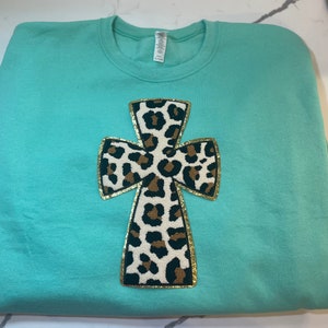 May include: A turquoise sweatshirt with a gold-trimmed cross made of a leopard print fabric.