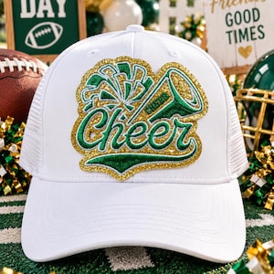 Può includere: Un cappellino trucker bianco con una toppa centrale ricamata in glitter oro e filo verde. La toppa mostra un megafono e un grafico a pon-pon con la parola Cheer scritta in un carattere corsivo al centro del design.