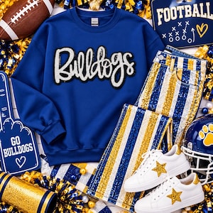 Puede incluir: Una sudadera azul real de cuello redondo con la palabra Bulldogs en letras blancas de lentejuelas, combinada con jeans de rayas de lentejuelas azules, doradas y blancas y zapatillas blancas con estrellas doradas, rodeada de accesorios de fútbol americano.