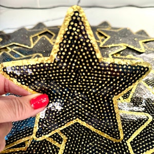 Op de afbeelding: Een zwarte stervormige patch met gouden pailletten en een gouden omtrek. De patch is gemaakt van stof en heeft een textuur.
