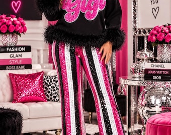 Peut inclure: Un sweat-shirt noir à manches longues avec une bordure en plumes noires au col et aux poignets, orné du mot Gigi en sequins roses sur la poitrine, associé à un pantalon taille haute à rayures verticales de sequins roses, noirs et blancs.
