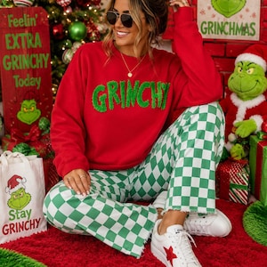 Könnte beinhalten: Ein rotes Langarm-Sweatshirt mit dem Wort GRINCHY in flauschigen grünen Buchstaben auf der Brust, kombiniert mit einer grün-weiß karierten Hose mit ausgefranstem Saum und weißen Sneakern mit einem roten Stern-Detail an der Seite.