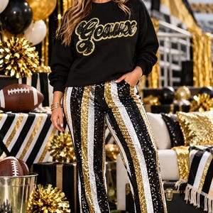 May include: A black long-sleeve sweatshirt featuring the word Geaux in gold sequin script lettering, paired with high-waisted trousers covered in vertical stripes of black, gold, and white sequins, finished with black block-heeled ankle boots.