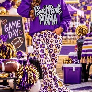 Könnte beinhalten: Ein violettes Langarm-Sweatshirt mit dem Text Ball Park MAMA in silberner Glitzerschrift, kombiniert mit einer weißen Hose mit violett-goldenem Leopardenmuster und weißen Turnschuhen mit einem violetten Stern-Detail an der Seite.
