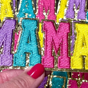 Peut inclure: Un ensemble de quatre patchs en feutre color&eacute;s &eacute;pellant le mot "MAMA" avec une bordure dor&eacute;e paillet&eacute;e. Chaque lettre est soulign&eacute;e d'or et a une couleur diff&eacute;rente : violet, turquoise, rose et jaune.