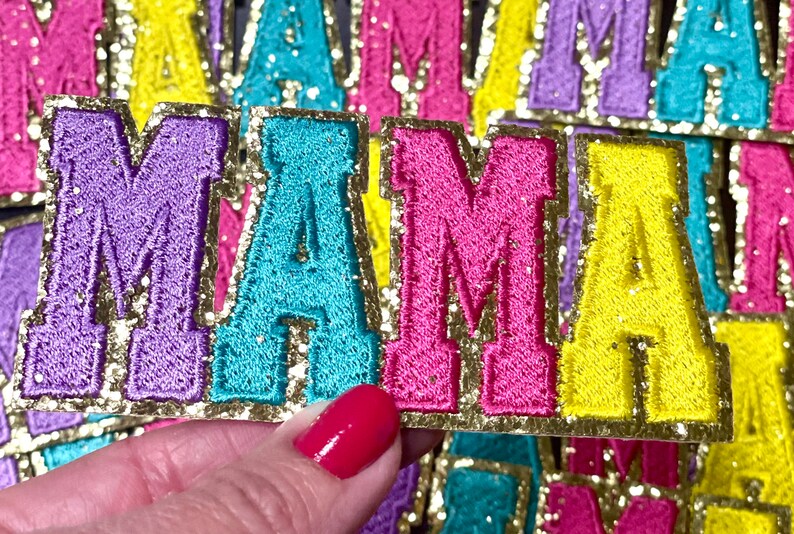 Peut inclure: Un ensemble de quatre patchs en feutre color&eacute;s &eacute;pellant le mot "MAMA" avec une bordure dor&eacute;e paillet&eacute;e. Chaque lettre est soulign&eacute;e d'or et a une couleur diff&eacute;rente : violet, turquoise, rose et jaune.