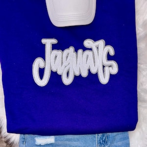 May include: A royal blue sweatshirt with the word "Jaguars" in a white, sequined script. A white baseball cap rests above the sweatshirt, and the bottom of the image shows a pair of light blue denim jeans.