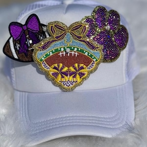 May include: White trucker hat adorned with three decorative patches. One patch is a football with a purple bow, another is a football-themed design, and the third is a purple paw print. All patches are embellished with glitter and pearls.