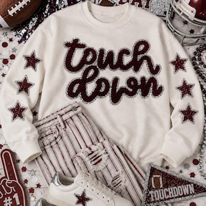 May include: A cream-colored crewneck sweatshirt featuring the text "touch down" in dark red glittery letters with silver outlines. Both sleeves are decorated with three dark red glittery stars. The sweatshirt is paired with striped pants.