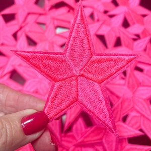 May include: Close-up of a bright pink embroidered star patch. The star has a five-point design with detailed stitching. A collection of similar star patches are in the background, creating a vibrant and eye-catching display. Ideal for crafting and embellishments.