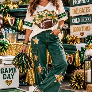 May include: A cream-colored V-neck t-shirt with green and gold sequined football and pom-pom graphics, paired with dark green wide-leg cargo pants featuring gold sequined star patches and white platform sneakers with metallic silver accents.