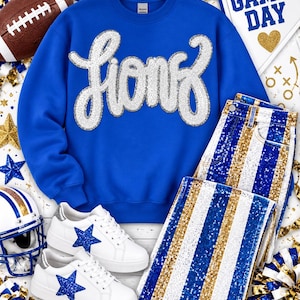 Puede incluir: Una sudadera azul real de cuello redondo con la palabra Lions en lentejuelas plateadas en el pecho, combinada con zapatillas blancas con estrellas de purpurina azul y pantalones con rayas verticales de lentejuelas azules, blancas y doradas.