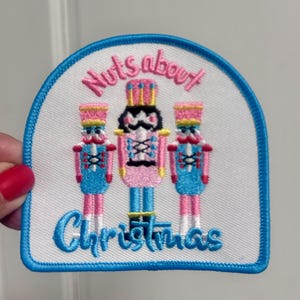 May include: A white iron-on patch with a blue border. The patch features three nutcracker figures in pink, blue, and yellow. The text "Nuts about Christmas" is written in a blue cursive font.