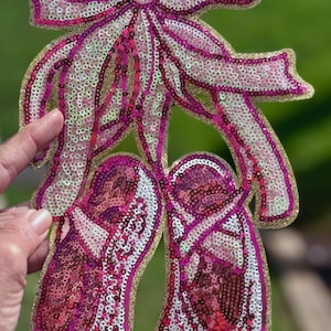 Op de afbeelding: Een paar roze balletten met een roze strik erop, allemaal gemaakt van pailletten. De schoenen zijn omrand met goud.