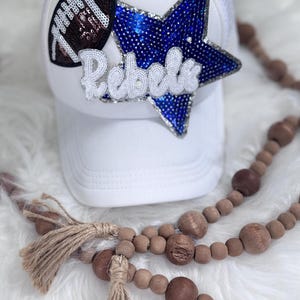 Puede incluir: Gorra de camionero blanca con un balón de fútbol y una estrella de lentejuelas azules. La palabra "Rebels" está escrita con lentejuelas blancas. Una guirnalda de cuentas de madera con borlas está en primer plano.
