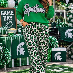 Puede incluir: Una sudadera verde brillante de manga larga con la palabra Spartans en letras de purpurina plateada en el pecho, combinada con pantalones de talle alto con estampado de leopardo negro, blanco y verde y zapatillas de plataforma blancas.