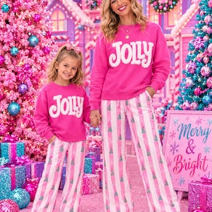 May include: Matching bright pink sweatshirts with white fuzzy lettering that reads JOLLY, paired with white and pink striped pajama pants featuring a small green Christmas tree pattern. The outfits are worn with white sneakers.