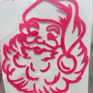Könnte beinhalten: Ein pinkfarbenes, strukturiertes Santa Claus-Gesicht-Design. Das Bild zeigt eine Nahaufnahme des Santa Claus-Gesichts, einschließlich Hut, Bart und rosigen Wangen. Das Design besteht aus einem flauschigen Material, das ihm ein weiches Aussehen verleiht.
