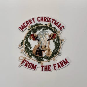 May include: White cow with brown spots in a wreath of green pine branches with gold stars. The wreath has the text "Merry Christmas from the Farm" in red.