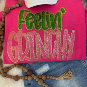 Puede incluir: Una sudadera rosa intenso con el texto "Feelin' Grinchy" en lentejuelas verdes y rosas. Una gorra de béisbol blanca, una guirnalda de cuentas de madera y unos pantalones cortos vaqueros también están en la imagen.