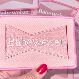May include: Pink satin patch with a white embroidered bow and the text "Babeweiser" in white.