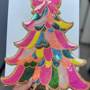 Puede incluir: Decoración de árbol de Navidad colorida con un lazo en la parte superior. El árbol está hecho de lentejuelas en tonos rosa, naranja, amarillo, verde y azul, delineado con un borde dorado. La decoración es sostenida por una mano.