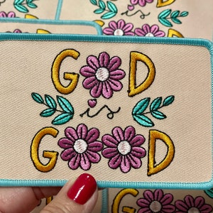 May include: A light blue embroidered patch with the text "God is Good" in yellow lettering. The text is surrounded by pink and green floral embroidery.