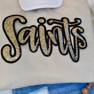 Op de afbeelding: Een beige sweatshirt met het woord "Saints" in gouden pailletten omlijnd met zwart. Een witte baseballpet is zichtbaar aan de bovenkant. De onderkant van de afbeelding toont een spijkerbroek.