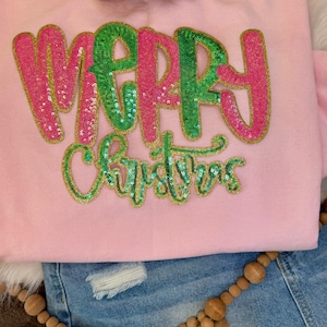 Może przedstawiać: Jasnoróżowa bluza z napisem "MERRY CHRISTMAS" w cekinowych literach. Słowo "MERRY" jest w różowych cekinach ze złotym obrysem, a słowo "CHRISTMAS" jest w zielonych cekinach ze złotym obrysem. Widoczna jest również biała czapka z daszkiem i niebieskie dżinsowe szorty.