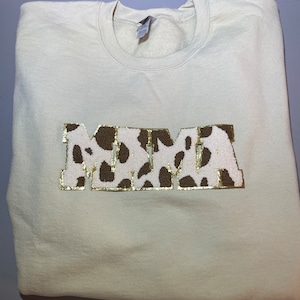 Puede incluir: Una sudadera de color crema con la palabra "MAMA" en letras grandes de estampado de leopardo marrón y blanco con detalles de purpurina dorada.