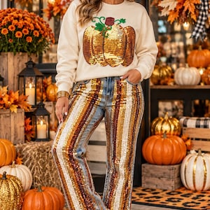 May include: A cream-colored long-sleeve sweatshirt featuring a large sequined pumpkin graphic on the front, paired with blue denim jeans adorned with vertical stripes of gold, bronze, and silver sequins, and white sneakers with a gold star detail.