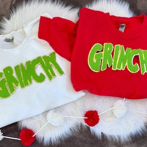 May include: Two crewneck sweatshirts, one white and one red, each with the word "GRINCHY" in large, green, sequined letters. A string of red and white pom-poms decorates the bottom. The sweatshirts are displayed on a white, fluffy surface.