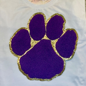 Könnte beinhalten: Lilafarbener Pfotenabdruck mit goldfarbenem Glitzerrand auf einem weißen T-Shirt. Der Pfotenabdruck besteht aus einem weichen, flauschigen Material.