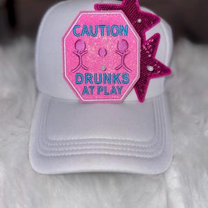 May include: White trucker hat with a pink and blue embroidered patch reading "CAUTION DRUNKS AT PLAY." Two sequined pink stars with pearl accents are attached. The hat has a curved peak and a mesh back.