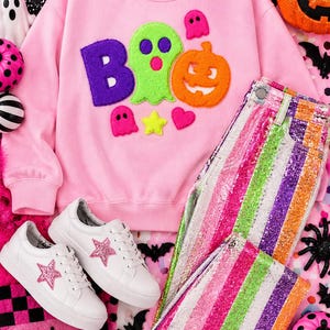 May include: A light pink crewneck sweatshirt featuring a large fuzzy applique that spells out BOO with a green ghost and orange pumpkin. Paired with white sneakers with pink glitter stars and pants featuring vertical stripes of pink, green, and purple glitter.