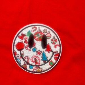 Puede incluir: Un parche blanco con forma de cara sonriente con un diseño navideño rojo, verde y azul. El parche está bordado con bastones de caramelo, adornos y árboles de Navidad.
