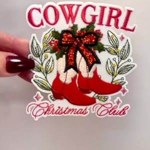 以下が含まれることがあります： 「Cowgirl Christmas Club」という文字と、赤いリボンと緑のホーリーが付いた赤いカウボーイブーツのペアが描かれた赤い刺繍パッチ