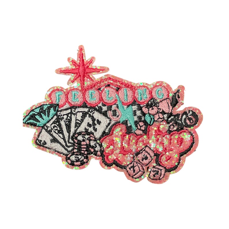 May include: Embroidered patch with a pink glitter border and the words "FEELIN' LUCKY" in a cursive font. The patch features playing cards, dice, and a starburst design, with accents of black, teal, and pink.