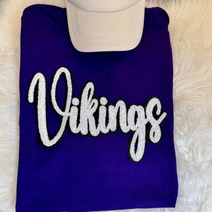 Może przedstawiać: Fioletowy t-shirt z napisem Vikings wykonanym białą kursywą z czarnym obrysem, umieszczony pod gładką białą czapką z daszkiem. Napis jest wykonany w teksturowanym stylu z cekinów na środku fioletowej tkaniny.