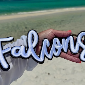 Può includere: Toppa ricamata blu chiaro e bianca con la scritta "Falcons" in corsivo. La toppa ha un bordo nero con dettagli glitterati. Lo sfondo è una scena di spiaggia con acqua turchese e sabbia bianca.