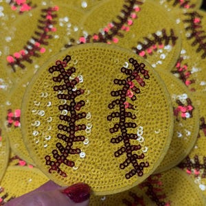 May include: Yellow sequined softball patches with red and pink stitching details. The patches are round and feature the classic softball seam design. The sequins add a sparkly, decorative element to the patches.